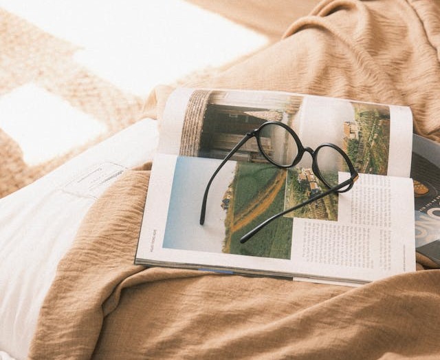 A cozy scene with a pair of black glasses resting on an open magazine, set atop a soft beige blanket in warm natural light.